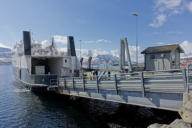 Bildet viser MF Helgøy, ved brygga i Hansnes, med Reinøya på andre siden av sundet. Bildet er tatt 6. mai 2019, en solrik dag på våren i Nord-Norge.
 Foto: Wolfmann, Wikimedia