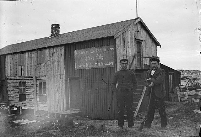 To menn fotografert foran "Jørgensens Etablissement. Kjøb og Salg. Fiskeprodukter - Handelsvarer". Bildet skal være tatt på Hansholmen i Sklinna i 1906.
 Foto: Anders Folkestadås/wikimedia commons
